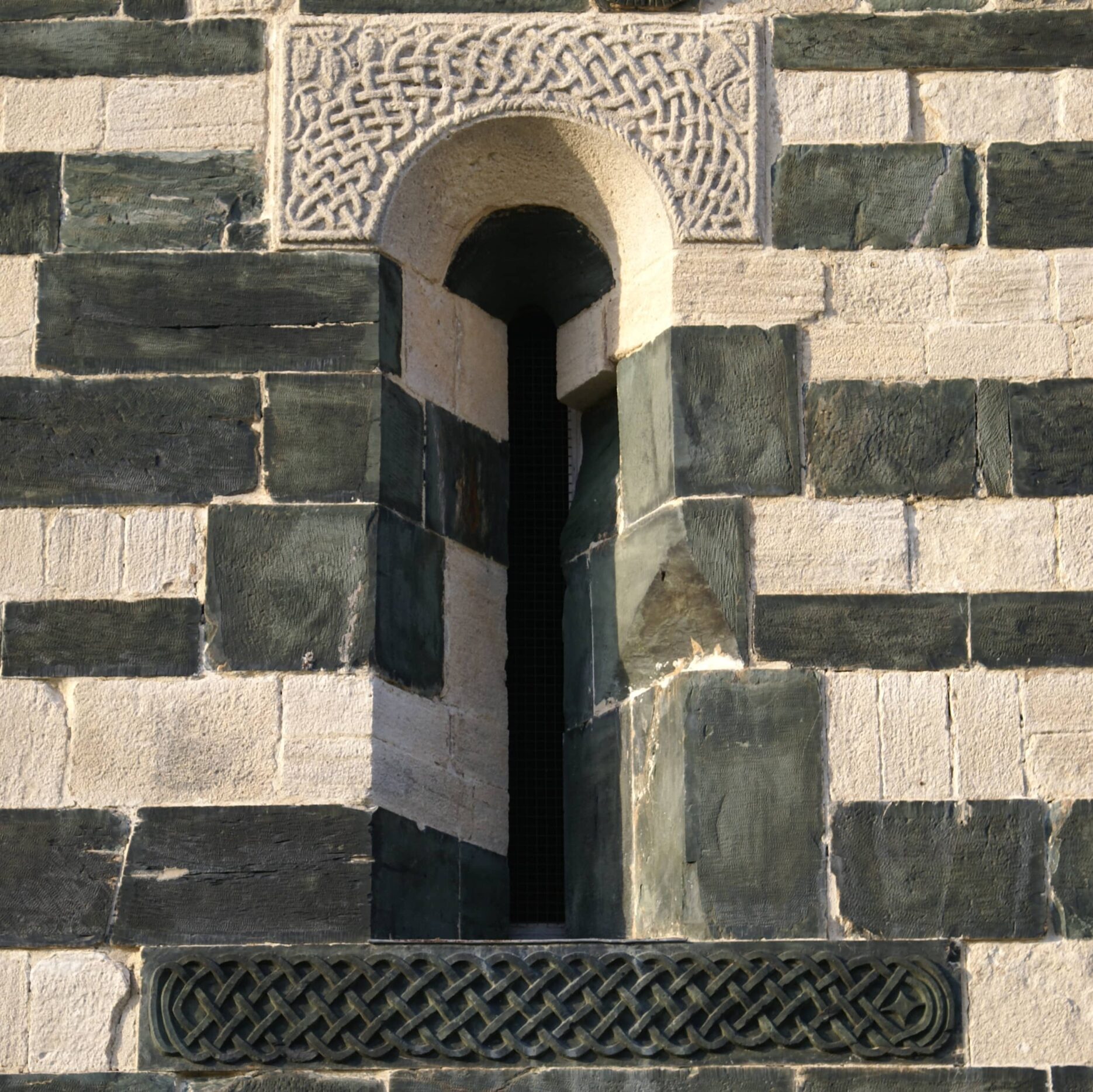 Une meurtrière dont le linteau et l'appui sont taillées de décorations : en haut des entrelacs entourés de vignes sur une pierre blanche calcaire, en bas un entrelacs caractéristique taillé dans une pierre verte de serpentinite.